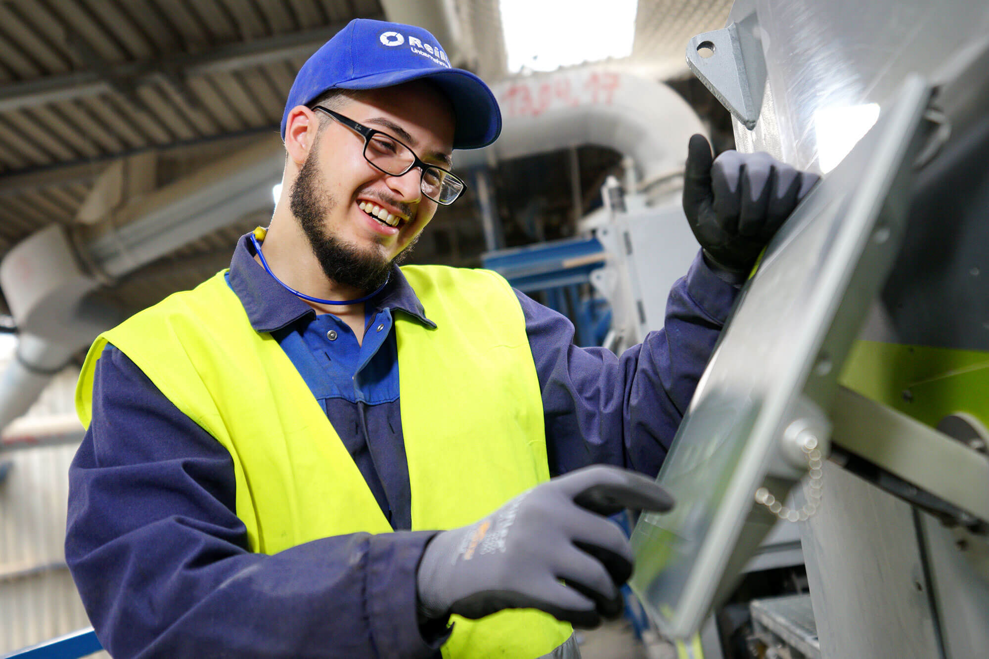 Maschinenfuehrer_reiling Ein junger Mann in seiner Ausbildung zum Maschinenführer bei der Firma Reilling. Mit sichtlichem Spaß bedient er die Maschinen auf der Arbeit. Er trägt stolz eine Reilling-Cap sowie die typische blaue Arbeitskleidung mit einer gelben Warnweste über dem Blaumann, die seine Sicherheit betont.