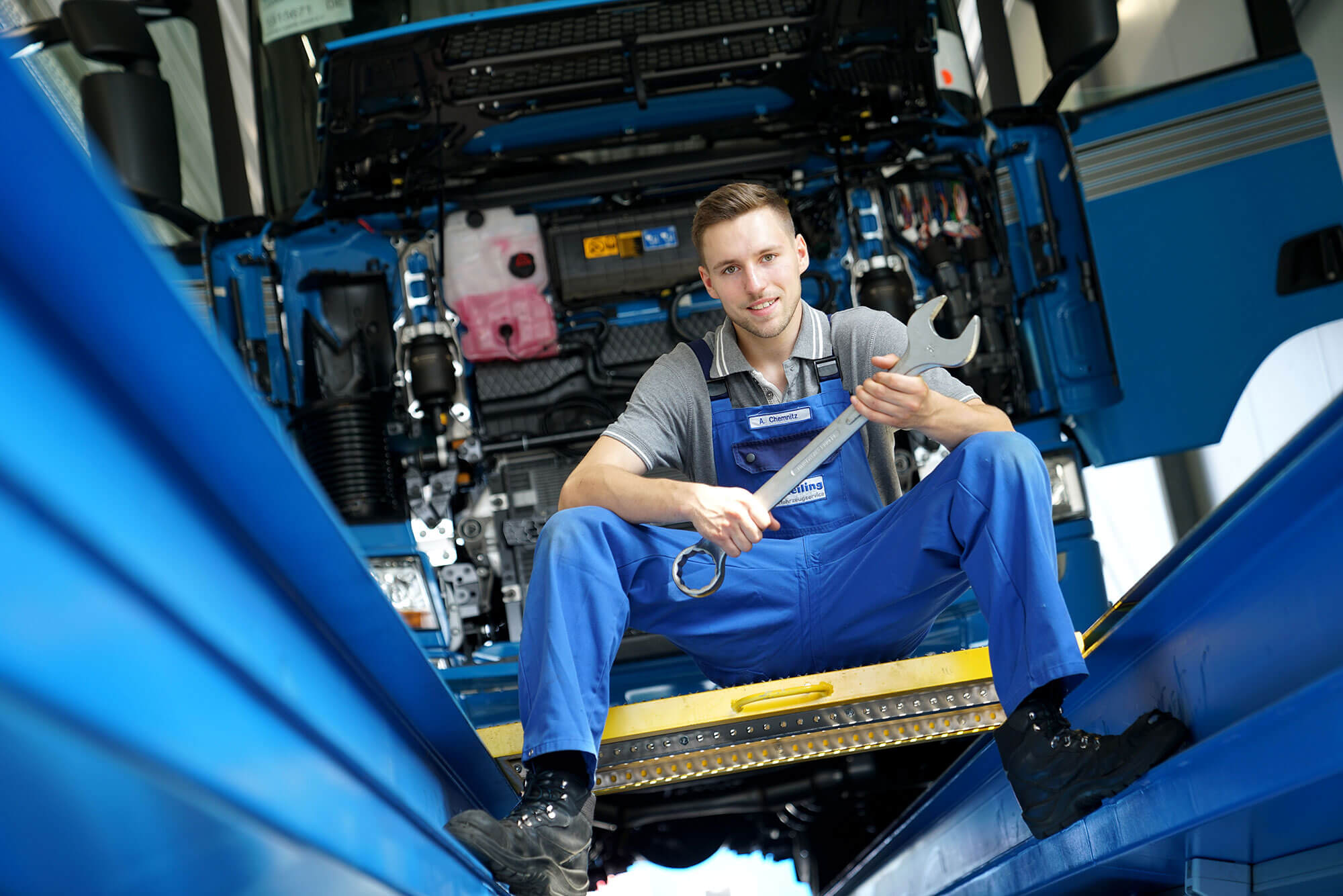 Mechatroniker_reiling Ein junger Mann in seiner Ausbildung bei Reilling als Mechatroniker für LKWs. Mit einem Schraubenschlüssel in der Hand sitzt er lächelnd an einer LKW-Fahrzeuggrube. Im Hintergrund ist ein großer LKW mit geöffneter Front zu erkennen, bereit, von ihm repariert zu werden.