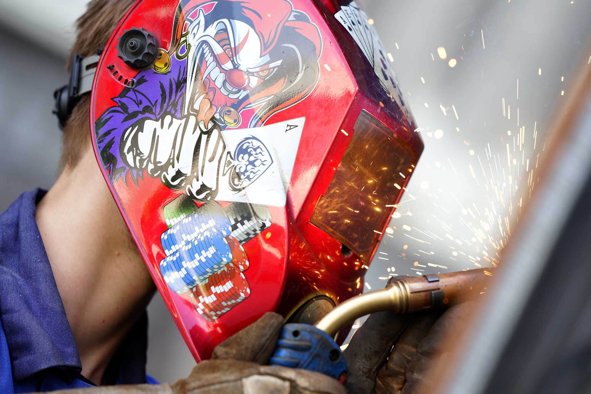 Ausbildung Konstruktionsmechaniker Reilling Ein junger Mann in seiner Ausbildung als Konstruktionsmechaniker bei der Firma Reilling. Er schweißt mit konzentriertem Blick, während die Funken fliegen. Auf seiner roten Schweißer-Maske sind Jetons zu sehen. Der Joker mit einer Pik-Ass-Spielkarte auf der Maske verleiht ihm einen einzigartigen Look.