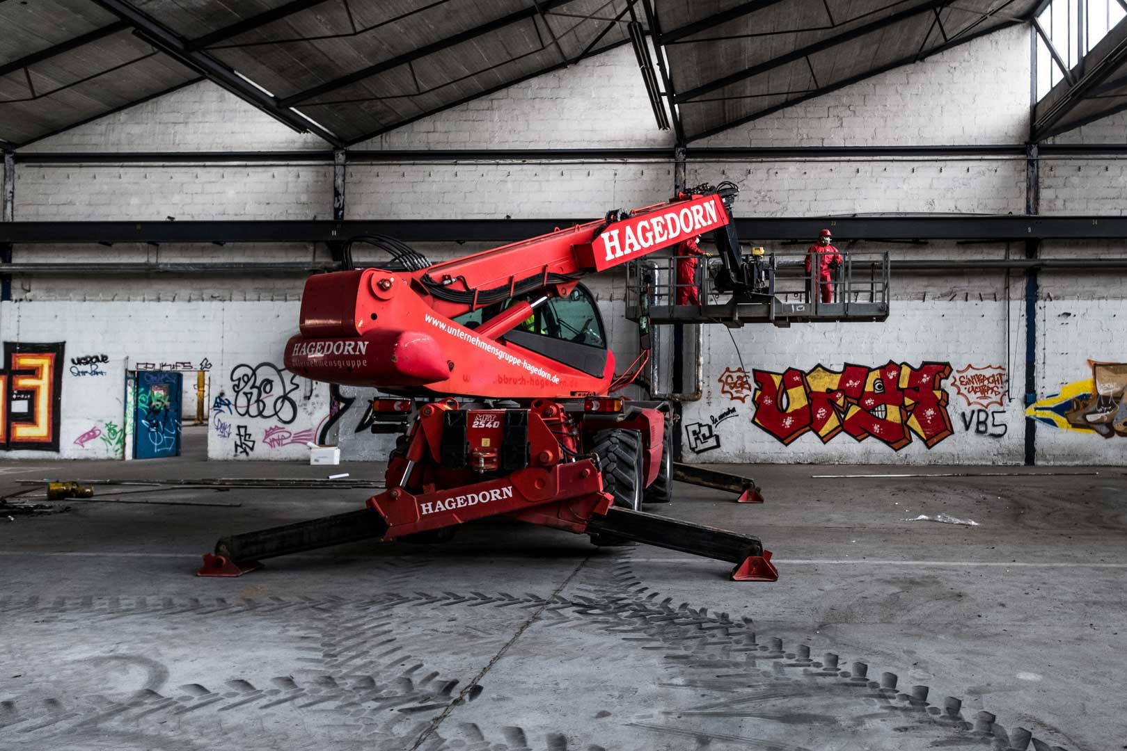 Hagedorn Hubsteiger In der geräumigen Halle mit kreativen Graffitis an den Wänden erstrahlt ein imposanter, roter Hubsteiger von Hagedorn. Mutig steht ein Arbeiter im Korb des Steigers, ausgestattet mit passender Arbeitskleidung und Helm, und genießt die faszinierende Perspektive von oben.