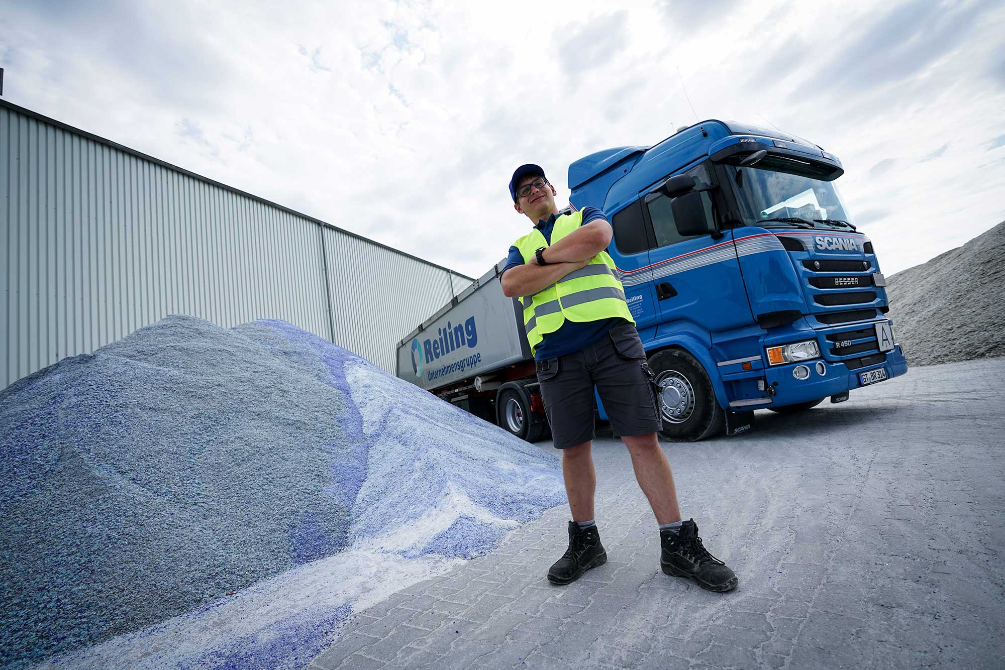 Ausbildung Berufskraftfahrer Reilling Ein junger Mann, stolz in seiner Ausbildung als Berufskraftfahrer bei der Firma Reilling. Er steht vor seinem blauen Scania LKW, während Schüttgüter für den Transport bereitstehen. Die graue Sattelauflieger mit dem Reilling-Aufdruck vervollständigt das Bild. Der Fahrer trägt eine gelbe Warnweste und strahlt Selbstbewusstsein aus