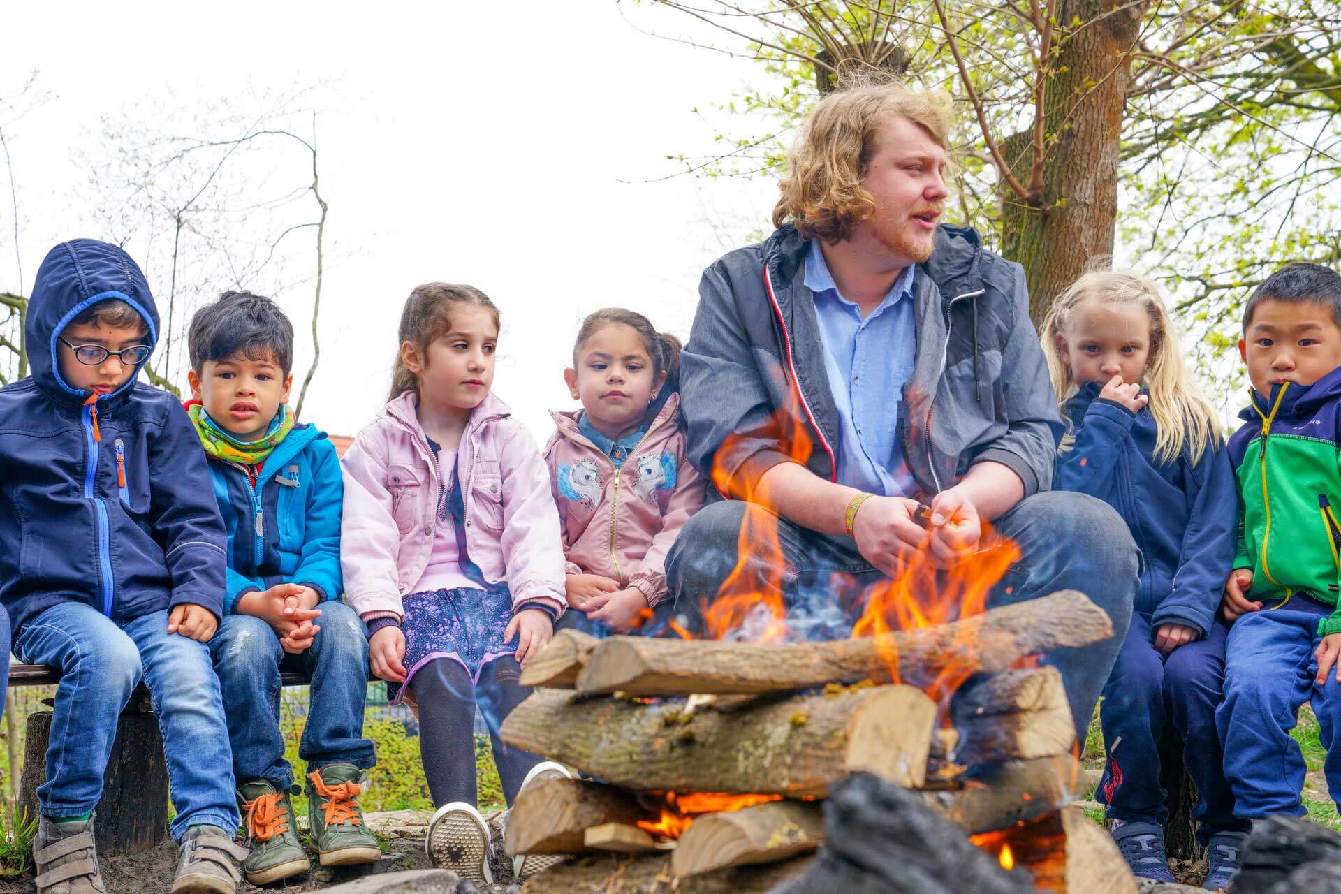 Gemeinschaft und Abenteuer: Lagerfeuerzeit mit unseren Kindern Ein Kindergärtner und fröhliche Kinder sitzen gemeinsam tagsüber um ein Lagerfeuer.