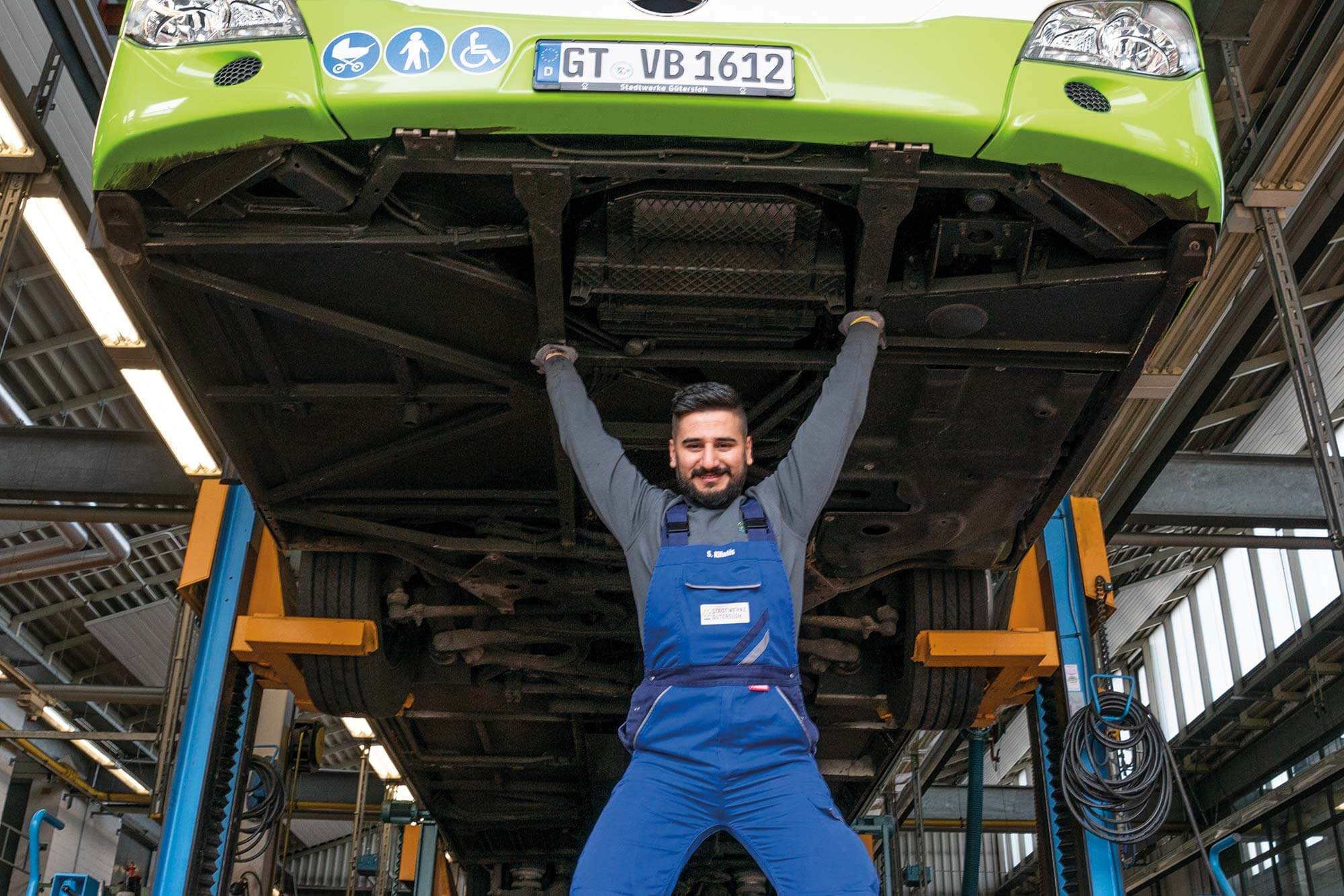 Schwere Arbeit leicht gemacht: Unser Mechatroniker zeigt seine Superkräfte in humorvoller Moment: Der Busmechatroniker scheint den ganzen Bus auf seinen Händen zu tragen, während das Fahrzeug auf einer Hebebühne steht.