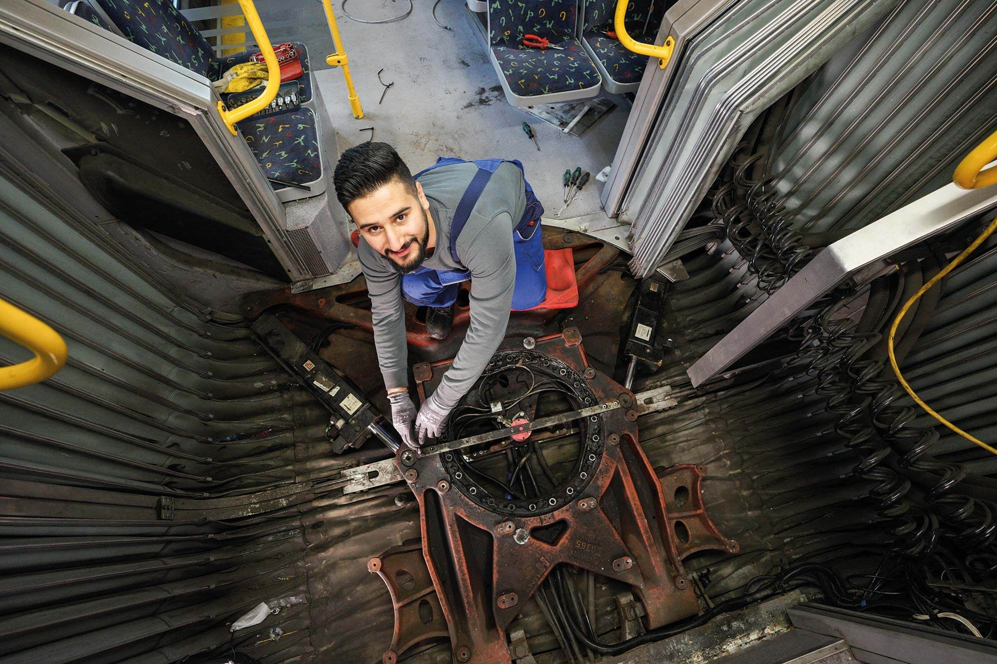 Fachmännische Reparatur: Unser leidenschaftlicher Mechatroniker in Aktion Ein junger Mitarbeiter der Stadt, ein begeisterter Mechatroniker für Busse, repariert das Gelenk eines Gelenkbusses mit einem glücklichen Lächeln.